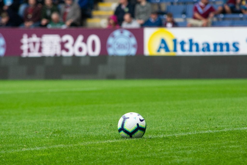 Fußball liegt auf grünem Rasen eines Premier-League-Stadions; im Hintergrund Tribüne und Fans.