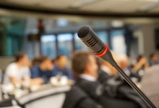 Ein Mikrofon an einem Rednerpult in einem mit Zuhörern besetzten Konferenzraum.